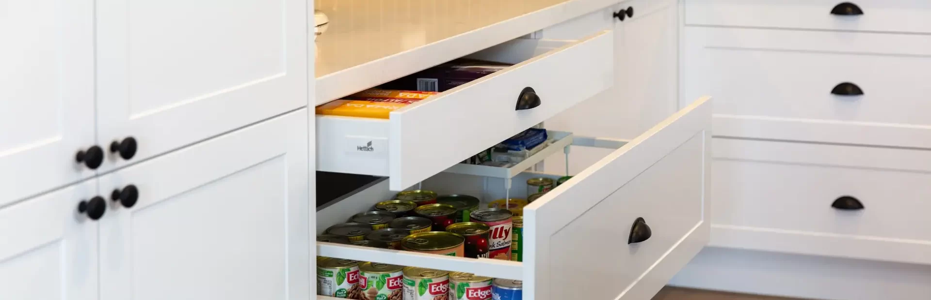 White shaker style kitchen drawer cabinets featuring tins of canned food and white stone benchtops with green tiled splashback