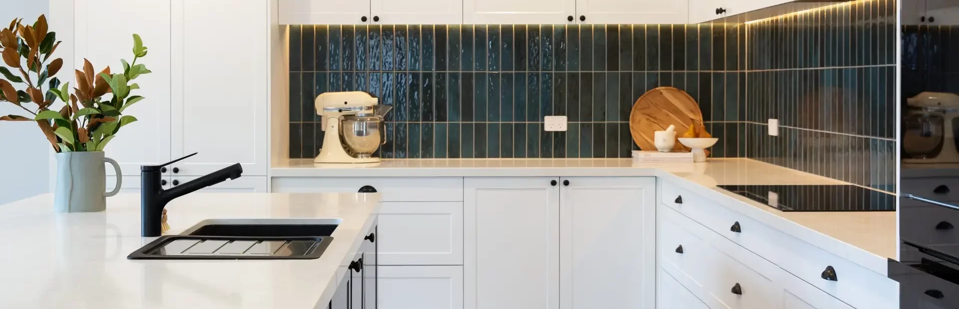 U shaped white and grey shaker style kitchen with white stone benchtops and green splashback tiling with styling items of boards and mix master