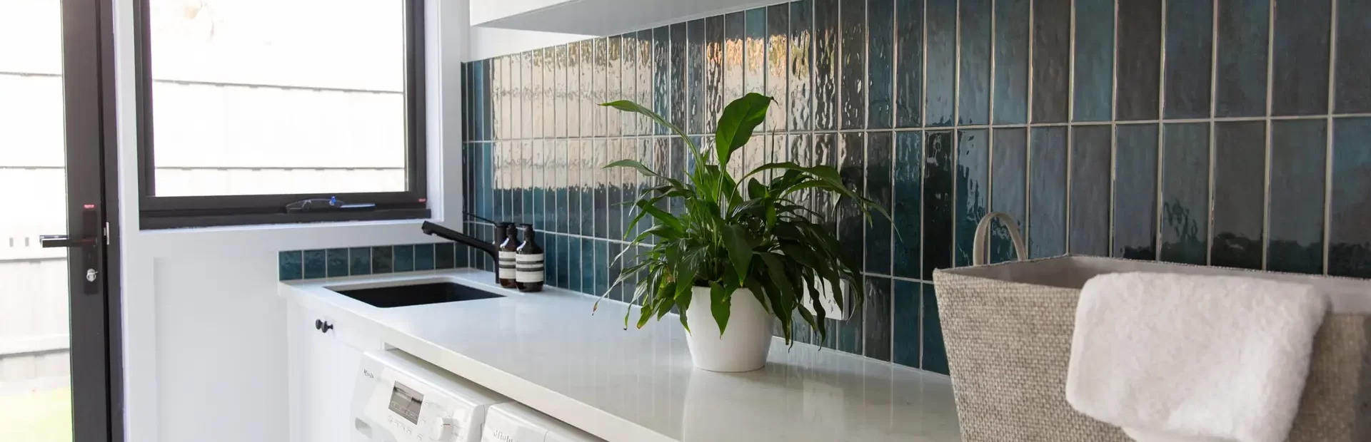 White kitchen cabinetry in galley style laundry with green splashback tiles leading to outside