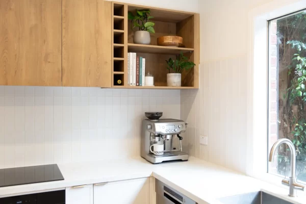 Corner kitchen with white hampton style cabinetry, white stone benchtops and oak coloured overhead cabinetry with cookbooks and open shelving displaying pot plants