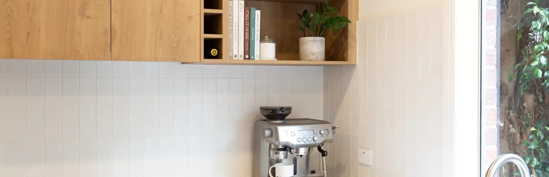 Corner kitchen with white hampton style cabinetry, white stone benchtops and oak coloured overhead cabinetry with cookbooks and open shelving displaying pot plants