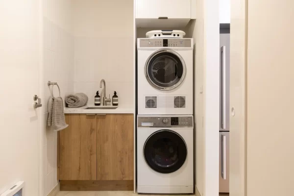 Compact laundry with washer and dryer stacked on top of each other and white overhead cabinet with slimline sink and oak underbench cabinetry