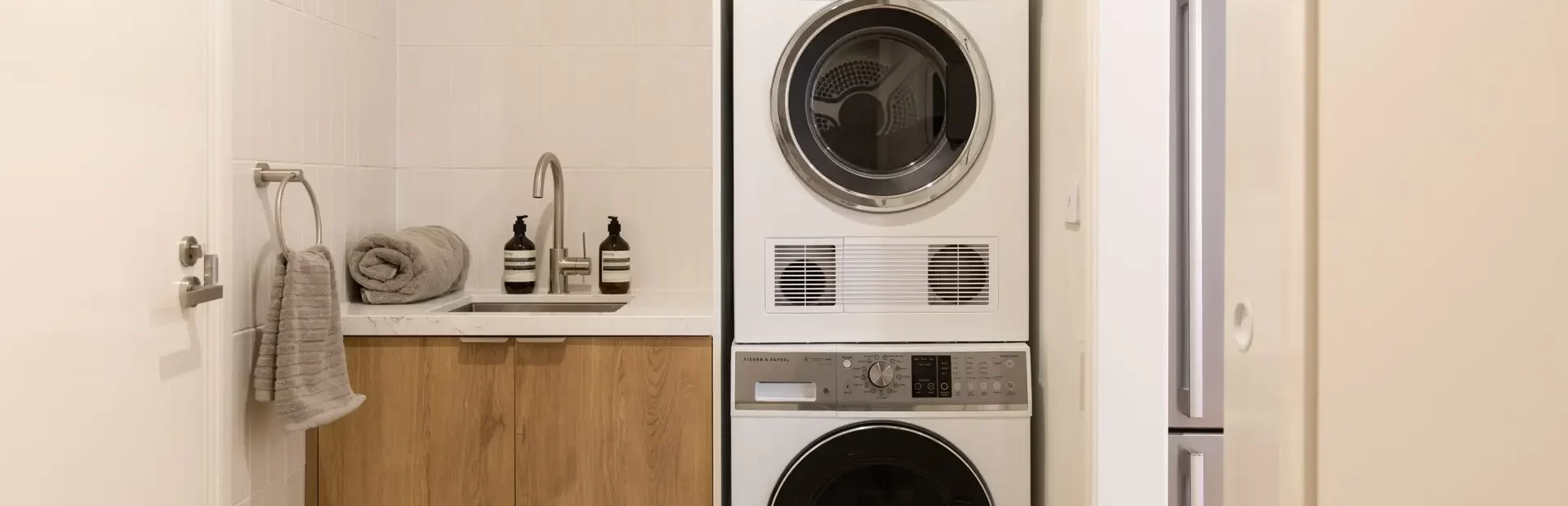 Compact laundry with washer and dryer stacked on top of each other and white overhead cabinet with slimline sink and oak underbench cabinetry