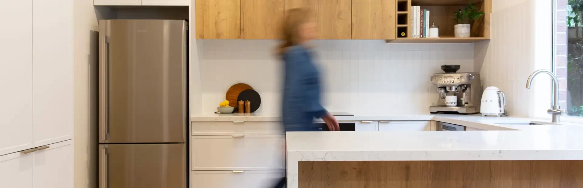 Female walking through galley kitchen with white kitchen cabinetry and oak timber look overhead cabinetry and island bench with white stone benchtops