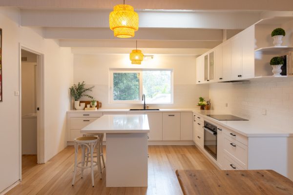 Modern white kitchen with shaker style cabinetry, timber flooring and central island bencht with stools. Features black oven and cooktop, open shelves with plants and two rattan pendant lights