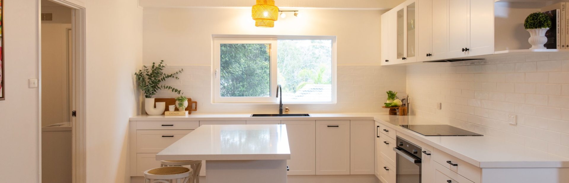 Modern white kitchen with shaker style cabinetry, timber flooring and central island bencht with stools. Features black oven and cooktop, open shelves with plants and two rattan pendant lights