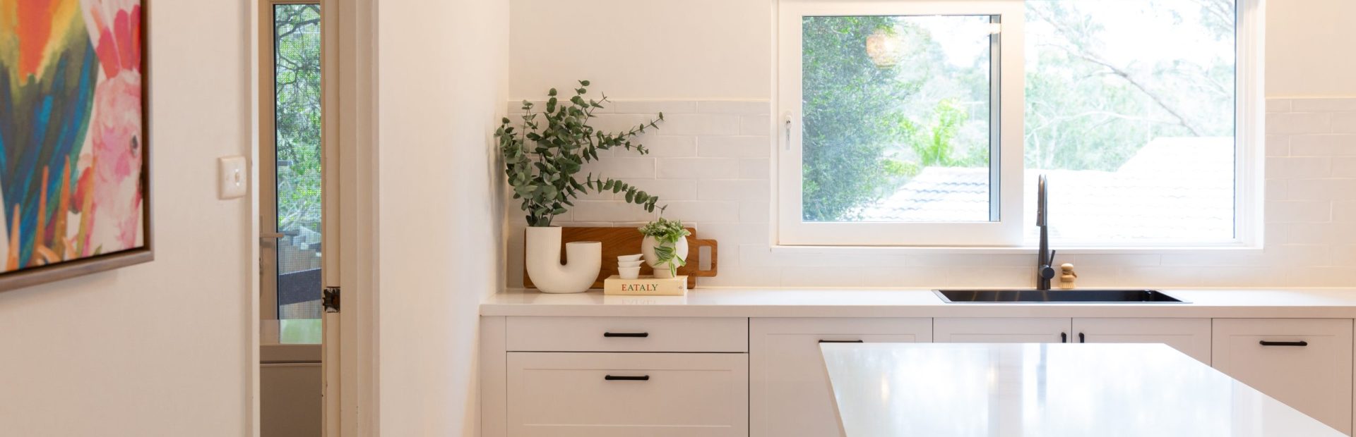 White kitchen cabinetry in shaker style with overhang central island bench and styling vase book and timber cutting board with greenery
