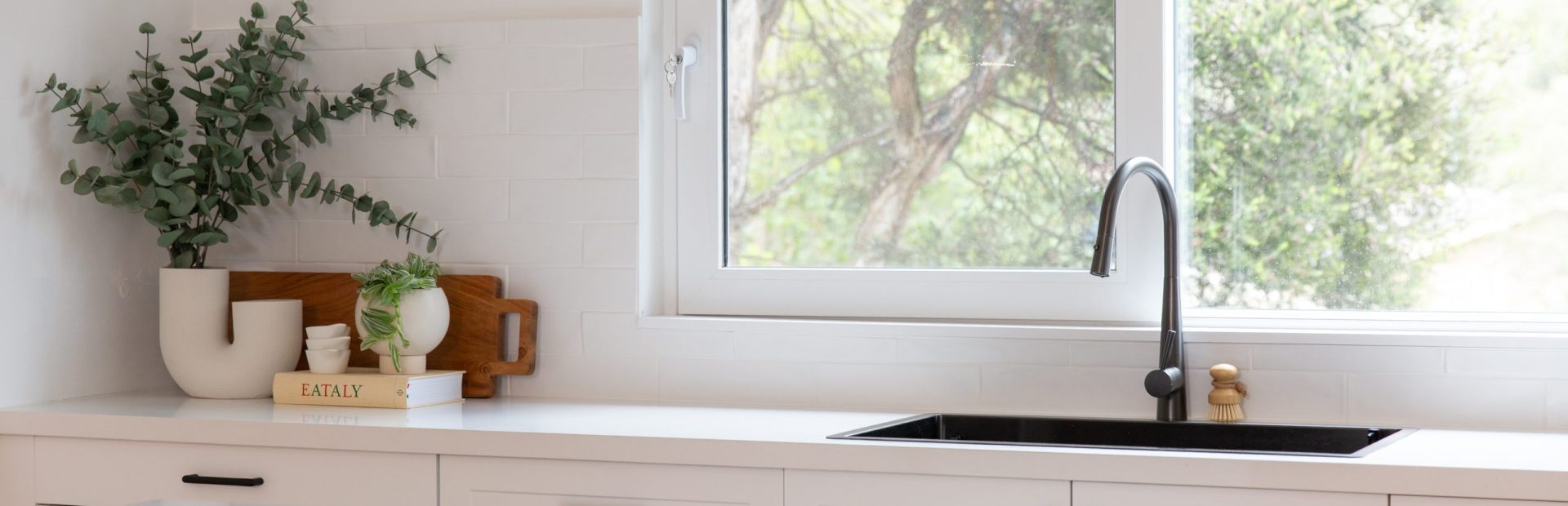 White shaker style kitchen cabinetry with black handles and black sink and tap with timber cutting board and pot plant
