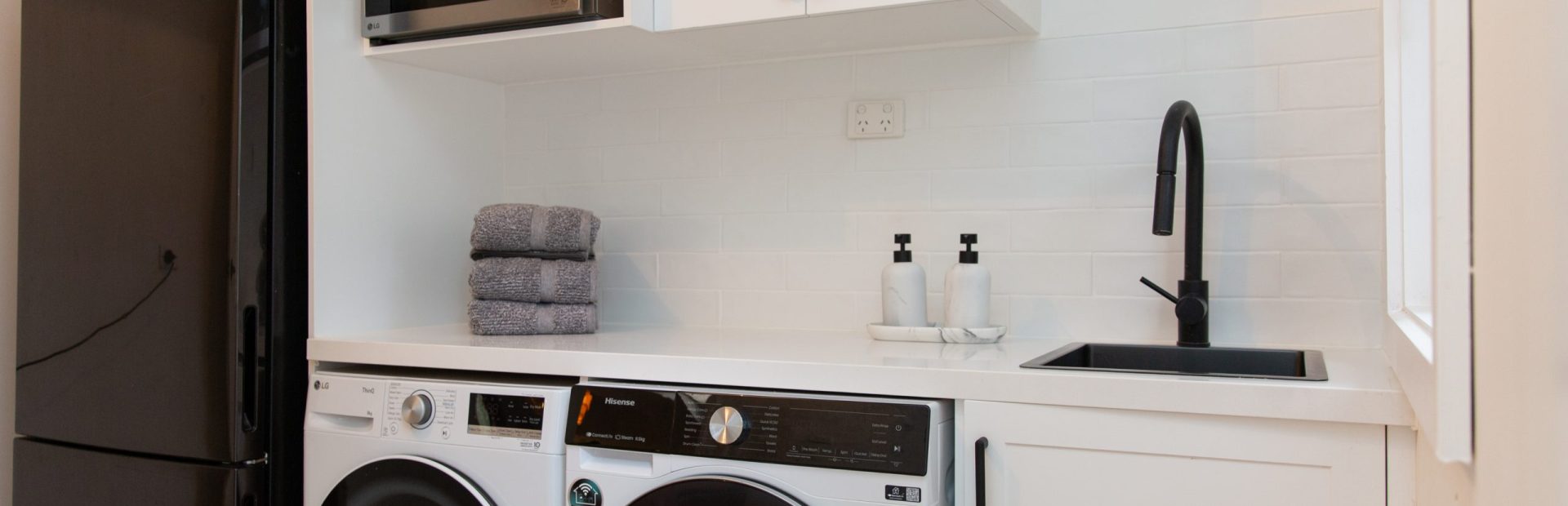 White shaker laundry cabinetry in galley shape with microwave cabinet and pile of grey towels with side by side washer and dryer