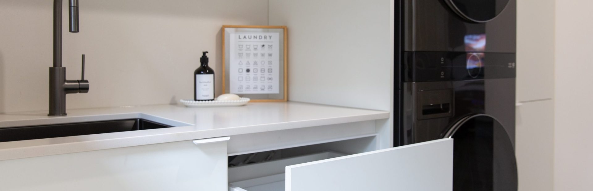 Galley laundry displaying white cabinet containing laundry hamper and double stacked black washer dryer combination