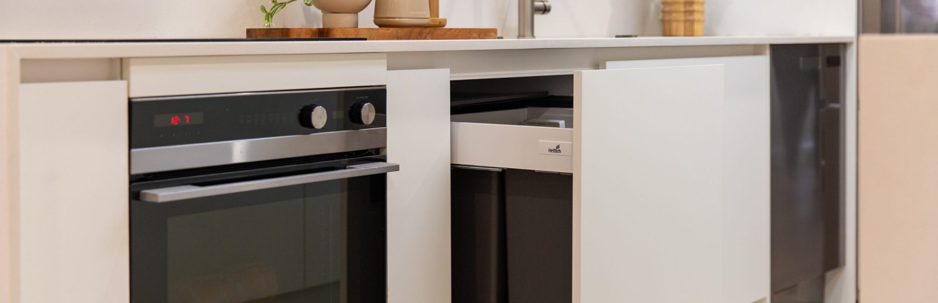 Kitchen displaying double pull out rubbish bins in white coloured cabinetry next to a black oven and plant and kitchen styling on benchtops