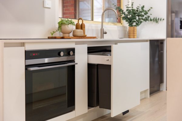 Kitchen displaying double pull out rubbish bins in white coloured cabinetry next to a black oven and plant and kitchen styling on benchtops