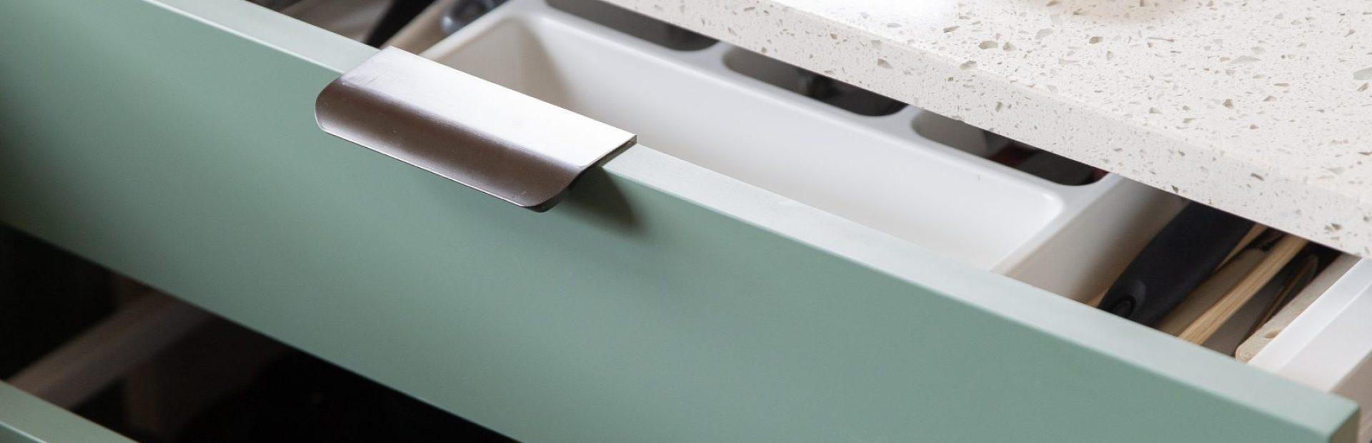 Green kitchen cabinet drawers with white cultery insert and brushed anthracite finger pull handles with white benchtops featuring wooden cutting board with slices of limes