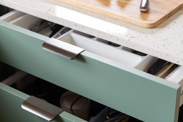 Green kitchen cabinet drawers with white cultery insert and brushed anthracite finger pull handles with white benchtops featuring wooden cutting board with slices of limes