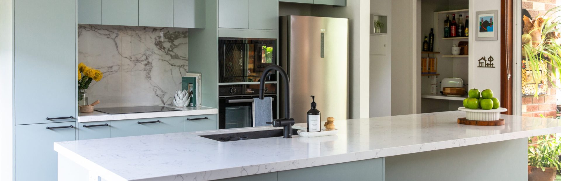 Green kitchen cabinetry with white stone benchtops featuring large standalone island bench